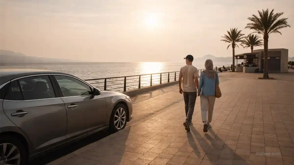 A couple walks along a Red Sea promenade in Saudi Arabia at sunset, with a silver sedan parked nearby and palm trees beside a small café kiosk.